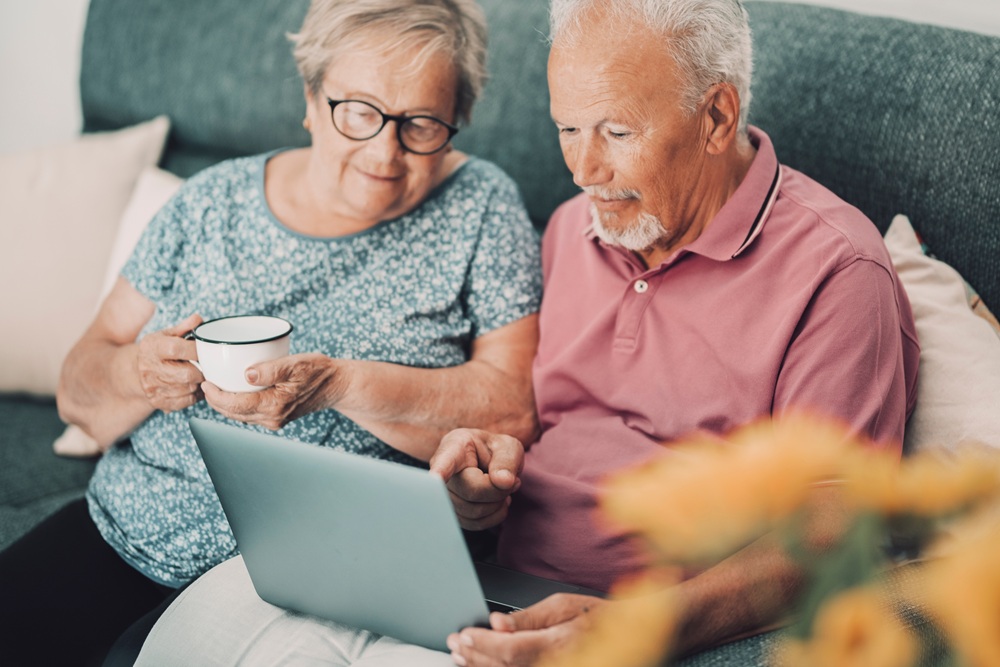 Elderly man and woman sitting closely on a sofa, smiling and looking at a laptop together—symbolizing digital independence and healthy vision with support from North Florida Eye Center